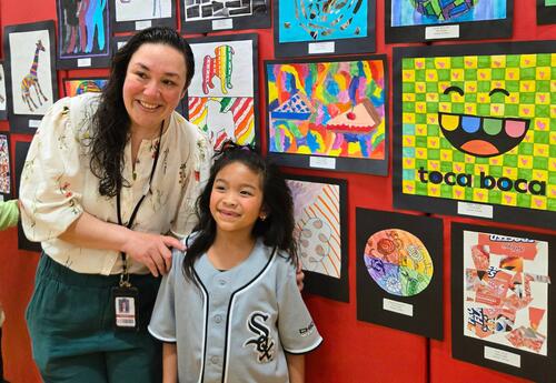 Art teacher Michelle Padilla and student posing in front of colorful animal-themed artwork displayed on a wall in a gallery setting.