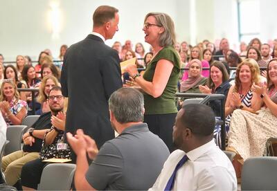 Two people standing, one receiving a recognition while a crowd sits and cheers.