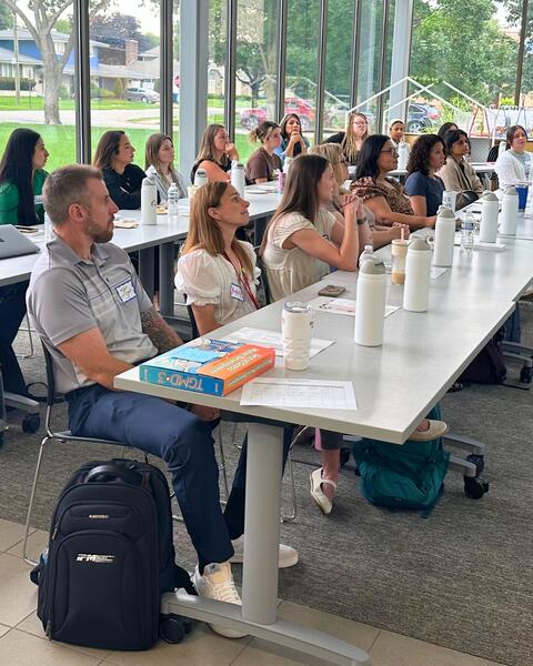 New staff members seated at long tables listening to a speaker standing.