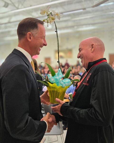 Two men smiling with a flower in front of a cheering crowd.