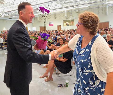 Two people shaking hands and one receiving a flower in front of a large, crowded room.