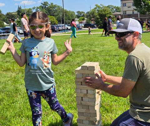 Young girl smiling and holding a wooden block while playing giant Jenga outdoors on a sunny day.