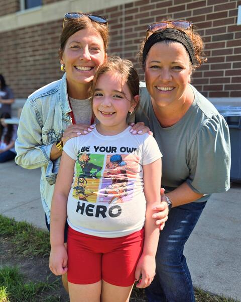 Smiling young girl in red shorts and a superhero-themed shirt stands between two women outdoors near a brick wall on a sunny day.