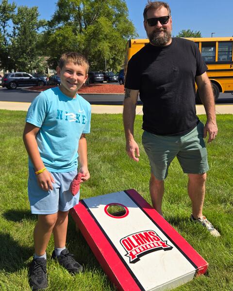 A boy and a man stand on grass next to a cornhole board with the Olhms Mustangs logo on a sunny day.