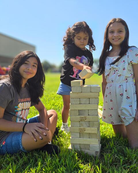Three girls playing a giant Jenga game outdoors on a sunny day with green grass and clear blue sky.