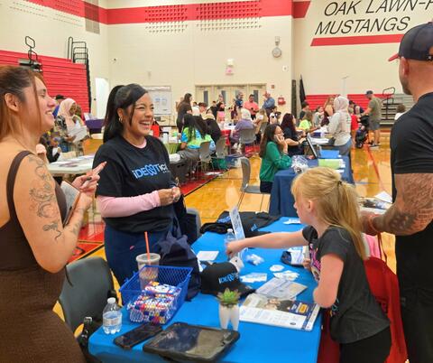 Volunteers at a community event table engaging with a young girl in a gymnasium with Oak Lawn-Mustangs signage.