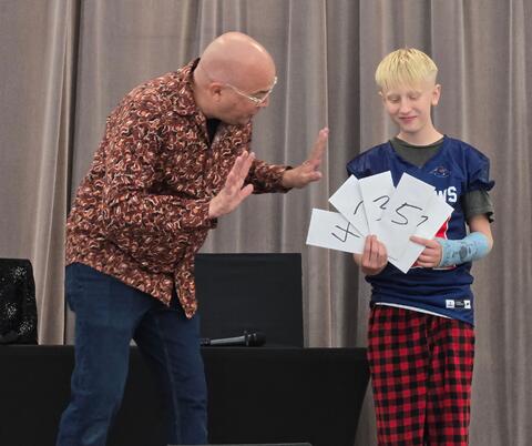 A man in a patterned shirt gestures excitedly toward a boy holding numbered cards during a stage performance.