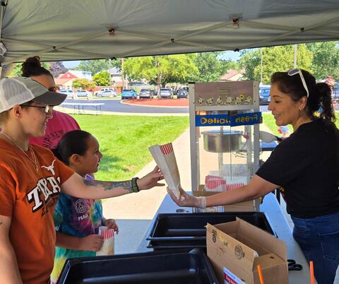 A woman serving popcorn to children under a canopy at an outdoor event on a sunny day.