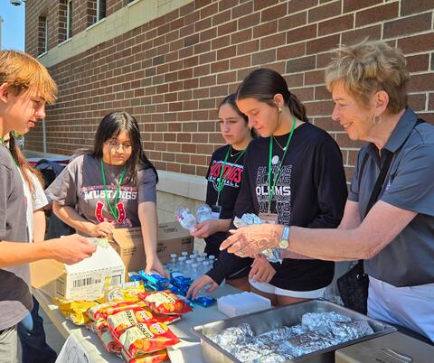 Group of young volunteers and an older woman distributing snacks and bottled water at an outdoor table against a brick wall.