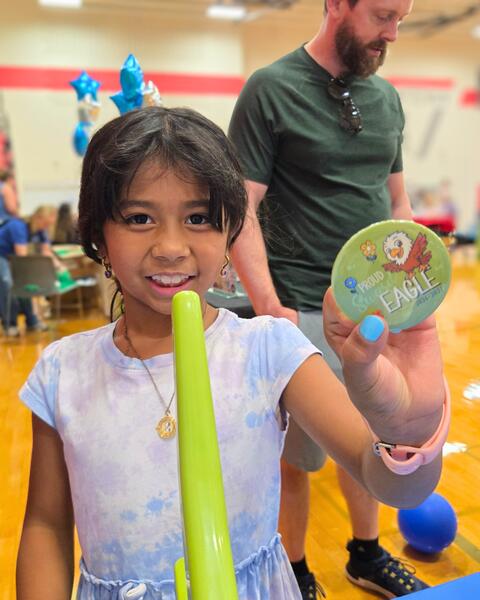 Smiling girl holding a green balloon sword and a 'Proud Eagle' badge in a gym with people in the background.