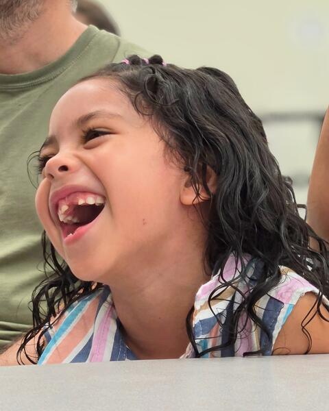 Young girl with curly hair laughing joyfully while sitting at a table indoors.