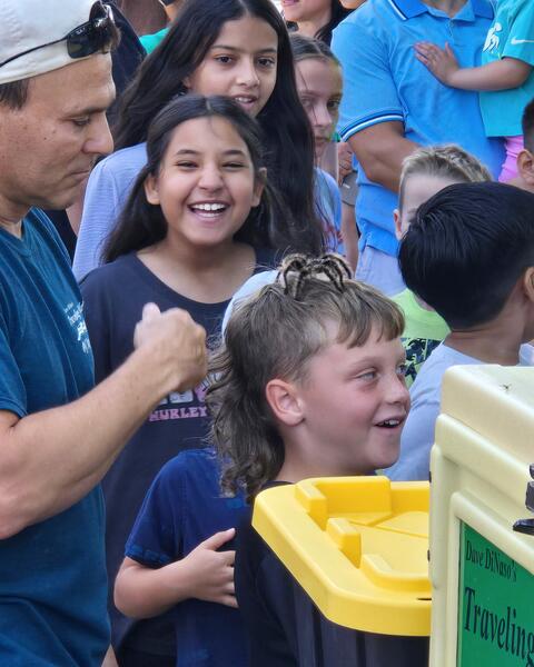 A group of smiling children in the background while a reptile vendor carefully oversees a tarantula on a young boy's head.
