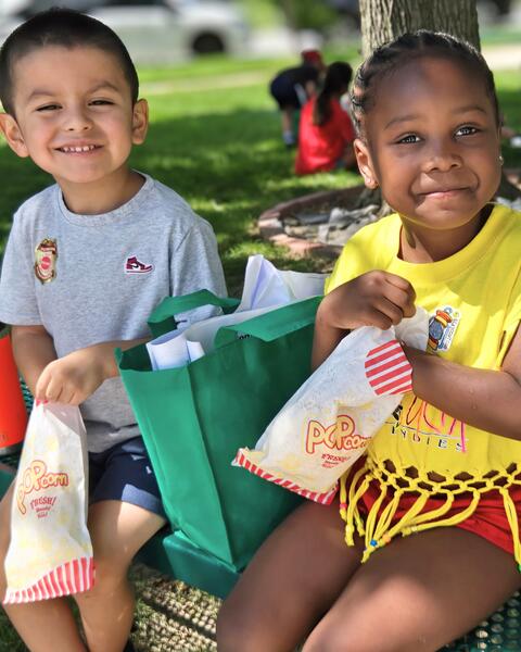 Two children sitting outdoors on a bench, each holding a bag of popcorn and smiling at the camera on a sunny day.