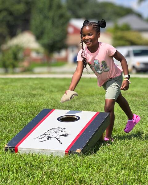Young girl smiling as she tosses a bean bag toward a cornhole board with a horse design on a sunny day outdoors.