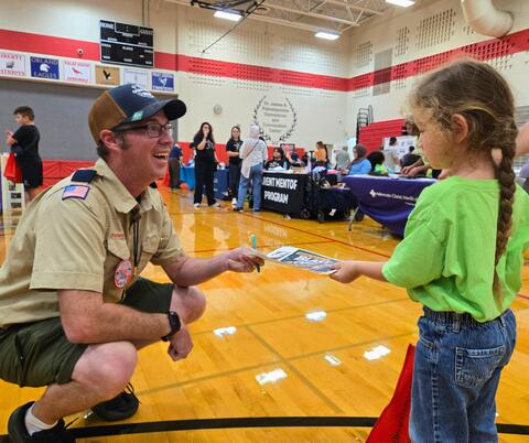 A scout leader kneels and smiles while handing a paper to a young girl in a green shirt inside a gymnasium event.