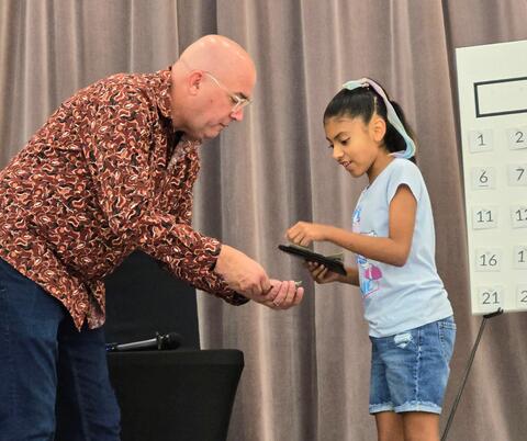 A man in a patterned shirt hands something to a young girl holding a large wallet during an indoor event.