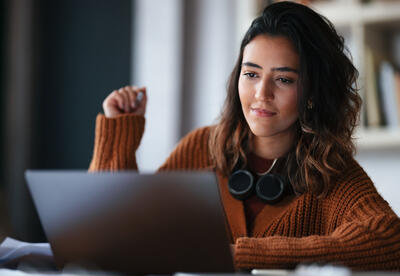 Here is a photo of a female student working on a laptop with headphones resting on her neck
