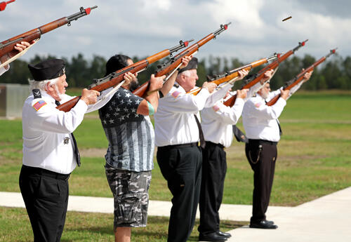 Veterans participate in a rifle salute during a Veterans Day ceremony, standing in a row outdoors and aiming rifles upward in honor of fallen service members.