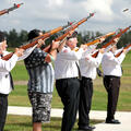 Veterans participate in a rifle salute during a Veterans Day ceremony, standing in a row outdoors and aiming rifles upward in honor of fallen service members.