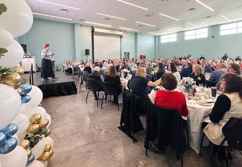 A photo: Chancellor Kristine Strickland speaks on stage during Fletcher Technical Community College’s Chancellor’s Breakfast. Attendees are seated at round tables throughout the room, and a balloon arrangement is positioned to the left of the stage.