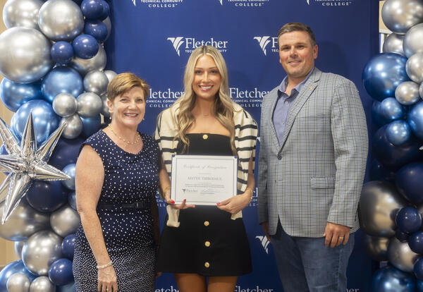 Three people stand in front of a Fletcher Technical Community College step-and-repeat backdrop with a blue and silver balloon arrangement. The student in the center holds a certificate of recognition, flanked by two college representatives, all smiling fo