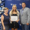 Three people stand in front of a Fletcher Technical Community College step-and-repeat backdrop with a blue and silver balloon arrangement. The student in the center holds a certificate of recognition, flanked by two college representatives, all smiling fo