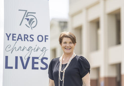 Fletcher Technical Community College Chancellor Dr. Kristine Strickland standing outdoors beside a sign that reads “75 Years of Changing Lives,” with campus buildings in the background.
