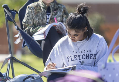 Two Fletcher Technical Community College students sit outside on campus studying. One student writes in a notebook while another reads a textbook from a folding chair. Backpacks rest on the grass nearby, with campus buildings softly blurred in the backgro