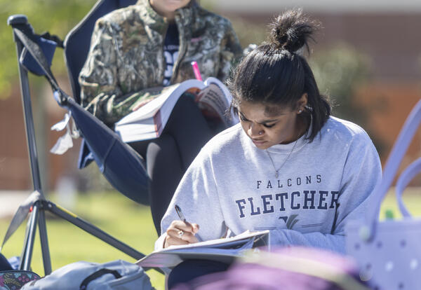 Two Fletcher Technical Community College students sit outside on campus studying. One student writes in a notebook while another reads a textbook from a folding chair. Backpacks rest on the grass nearby, with campus buildings softly blurred in the backgro