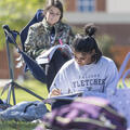 Two Fletcher Technical Community College students sit outside on campus studying. One student writes in a notebook while another reads a textbook from a folding chair. Backpacks rest on the grass nearby, with campus buildings softly blurred in the backgro