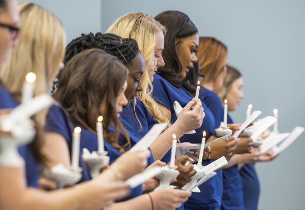 A line of Practical Nursing graduates in blue scrubs hold lit nursing candles and printed programs while reciting the Nightingale Pledge during Fletcher Technical Community College’s Nursing Pinning Ceremony.