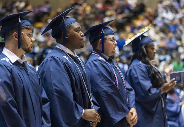 Fletcher Technical Community College graduates stand during the Spring 2026 Commencement Ceremony wearing blue caps and gowns. Several graduates are shown in profile as they listen attentively during the ceremony, with family members, friends, and support
