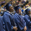 Fletcher Technical Community College graduates stand during the Spring 2026 Commencement Ceremony wearing blue caps and gowns. Several graduates are shown in profile as they listen attentively during the ceremony, with family members, friends, and support