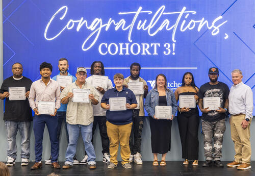 Eleven people stand on stage in front of a large blue screen reading "Congratulations, Cohort 3!" Ten graduates hold their Commercial Driver's License (CDL) certificates after completing Fletcher Technical Community College's CDL program through Venture G
