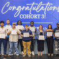 Eleven people stand on stage in front of a large blue screen reading "Congratulations, Cohort 3!" Ten graduates hold their Commercial Driver's License (CDL) certificates after completing Fletcher Technical Community College's CDL program through Venture G