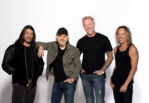 The four members of Metallica stand together in front of a white backdrop, smiling for a promotional photo. They are dressed casually in dark shirts and jeans, with one member wearing a black cap and another wearing a black leather jacket.