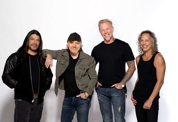 The four members of Metallica stand together in front of a white backdrop, smiling for a promotional photo. They are dressed casually in dark shirts and jeans, with one member wearing a black cap and another wearing a black leather jacket.