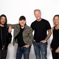 The four members of Metallica stand together in front of a white backdrop, smiling for a promotional photo. They are dressed casually in dark shirts and jeans, with one member wearing a black cap and another wearing a black leather jacket.