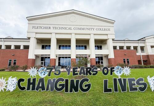 The front of Fletcher Technical Community College's Schriever campus is shown beneath a cloudy sky. On the front lawn, a colorful yard display with large bubble letters reads "75 Years of Changing Lives" and features blue, white, and silver starburst and 