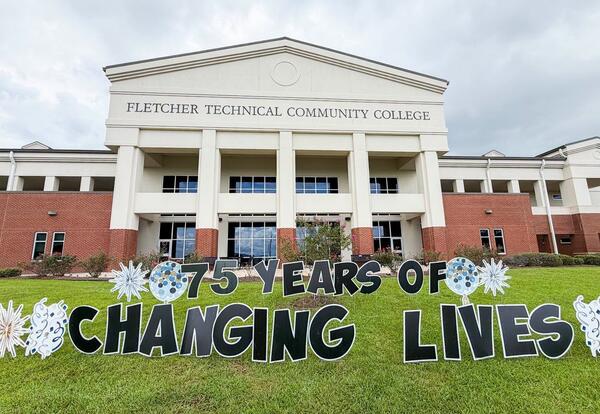 The front of Fletcher Technical Community College's Schriever campus is shown beneath a cloudy sky. On the front lawn, a colorful yard display with large bubble letters reads "75 Years of Changing Lives" and features blue, white, and silver starburst and 