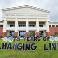 The front of Fletcher Technical Community College's Schriever campus is shown beneath a cloudy sky. On the front lawn, a colorful yard display with large bubble letters reads "75 Years of Changing Lives" and features blue, white, and silver starburst and 
