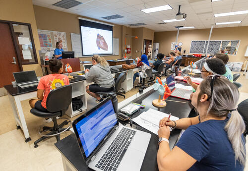 Biology Professor Erica Burns teaches a classroom of students using a projected diagram of the human heart in a biology laboratory. Students seated at lab stations follow along with laptops, notebooks, and heart models on their desks. Anatomical posters, 