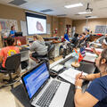 Biology Professor Erica Burns teaches a classroom of students using a projected diagram of the human heart in a biology laboratory. Students seated at lab stations follow along with laptops, notebooks, and heart models on their desks. Anatomical posters, 