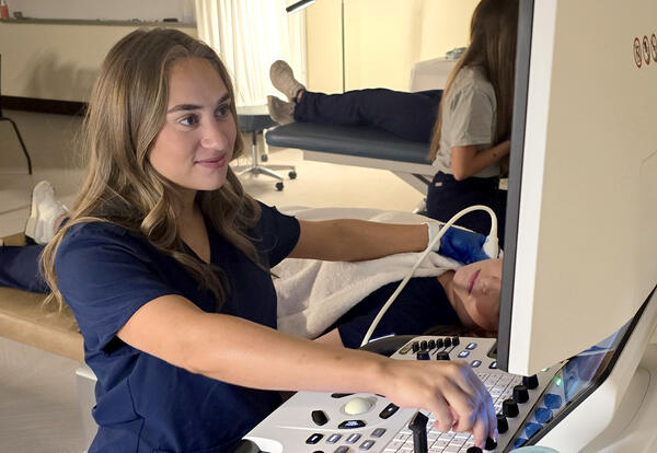 A Cardiovascular Sonography student in navy scrubs operates an ultrasound machine while scanning a patient during a hands-on lab exercise. Another student practices in the background.