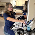 A Cardiovascular Sonography student in navy scrubs operates an ultrasound machine while scanning a patient during a hands-on lab exercise. Another student practices in the background.