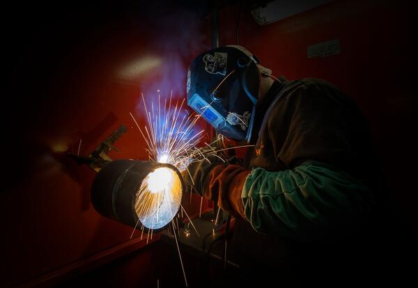A student wearing a protective welding helmet and safety gear welds a thick metal pipe, creating a bright burst of blue light and flying orange sparks inside a dark training booth.