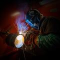 A student wearing a protective welding helmet and safety gear welds a thick metal pipe, creating a bright burst of blue light and flying orange sparks inside a dark training booth.