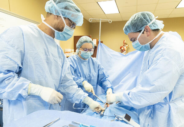 Fletcher Surgical Technology students wearing surgical gowns, masks and gloves practice a simulated procedure around an operating table in a healthcare training lab.