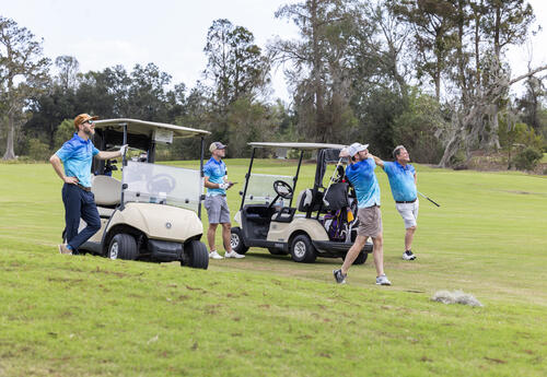 Four golfers wearing matching blue shirts stand near two golf carts as one golfer takes a swing on the course at Ellendale Country Club in Houma.