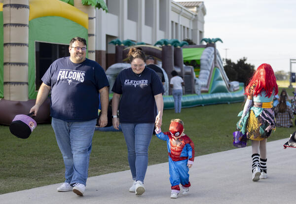 Trey and Josie Clark walk beside their young son, Andy, who wears a Spider-Man costume and holds Josie’s hand. Bounce houses and other costumed attendees are behind them at Fletcher’s Fall Fest.